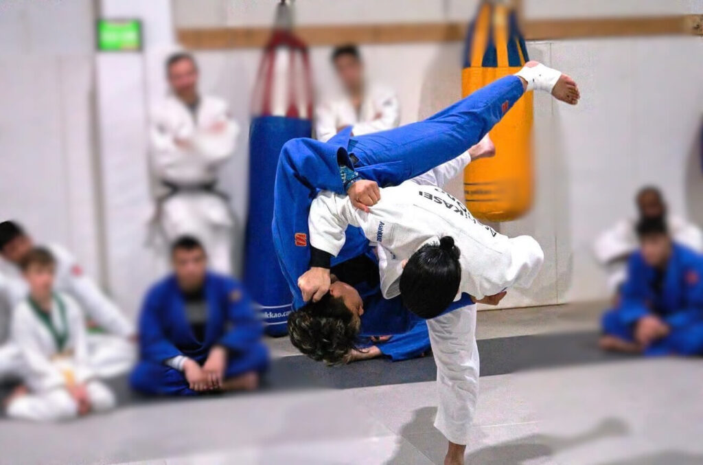 Spectators in a dojo watching as a judoka in a white gi throws an opponent in a blue gi at The Compound Classic Judo Shiai.