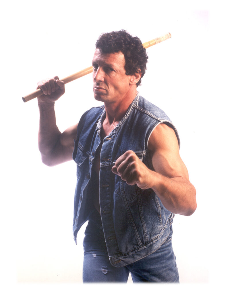 Punong Guro Marc 'Crafty Dog' Denny wearing a denim jacket and jeans, holding his right fist forward while swinging a rattan stick with his left hand.